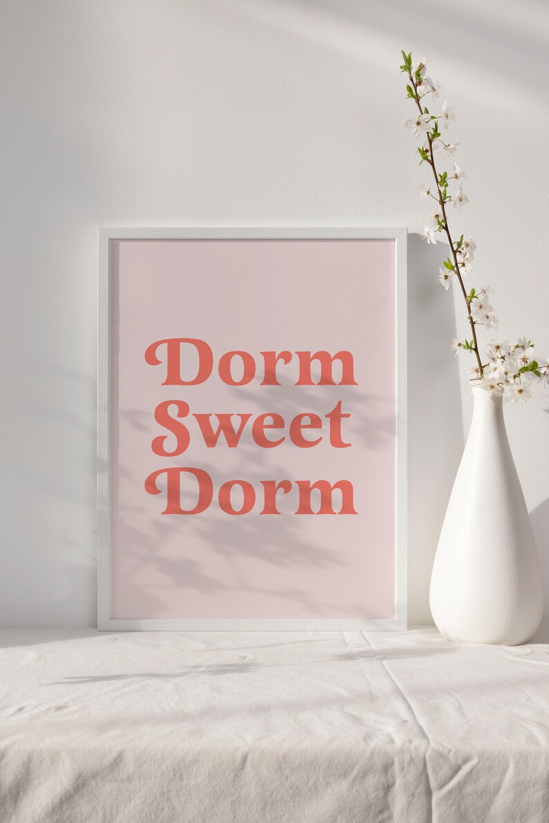 May include: A framed print with the words "Dorm Sweet Dorm" in coral-colored, retro-style lettering on a light pink background. A white vase with white flowers sits to the right of the framed print. The print is on a white surface.