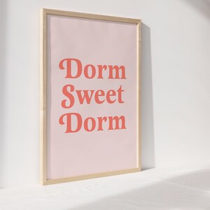 May include: A framed print with the words "Dorm Sweet Dorm" in coral-colored letters on a light pink background. The frame is made of light-colored wood. The print is displayed on a white surface.