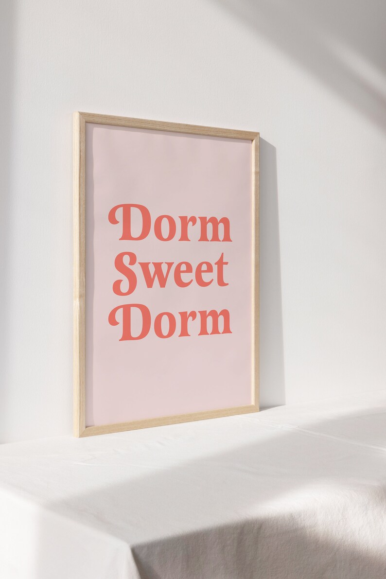 May include: A framed print with the words "Dorm Sweet Dorm" in coral-colored letters on a light pink background. The frame is made of light-colored wood. The print is displayed on a white surface.