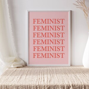 Puede incluir: Un estampado rosa enmarcado con la palabra "FEMINISTA" repetida en letras color coral. El estampado se muestra sobre una superficie beige texturizada, con un jarr&oacute;n blanco que contiene flores secas a la derecha. Una cortina blanca est&aacute; a la izquierda.