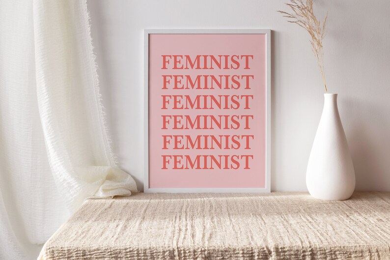 Puede incluir: Un estampado rosa enmarcado con la palabra "FEMINISTA" repetida en letras color coral. El estampado se muestra sobre una superficie beige texturizada, con un jarr&oacute;n blanco que contiene flores secas a la derecha. Una cortina blanca est&aacute; a la izquierda.