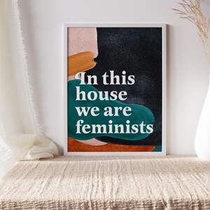 May include: A framed print with an abstract design in shades of orange, green, and black. The text "In this house we are feminists" is printed in white on the print.
