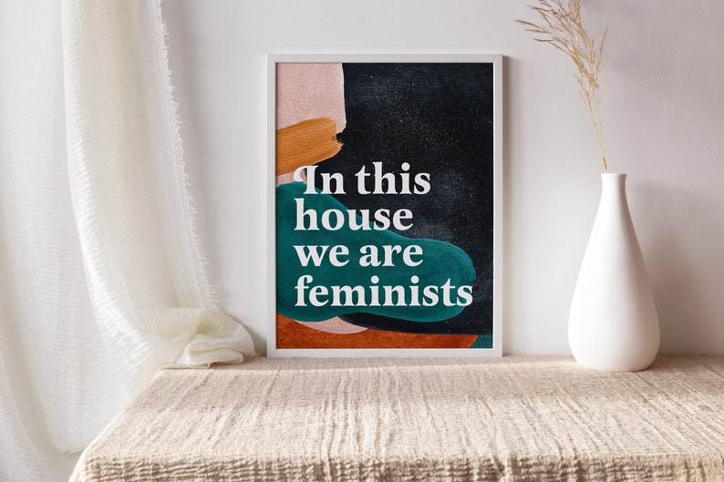 May include: A framed print with an abstract design in shades of orange, green, and black. The text "In this house we are feminists" is printed in white on the print.