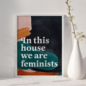 May include: A framed print with an abstract background in shades of pink, orange, brown, and teal. The text "In this house we are feminists" is printed in white on the print.