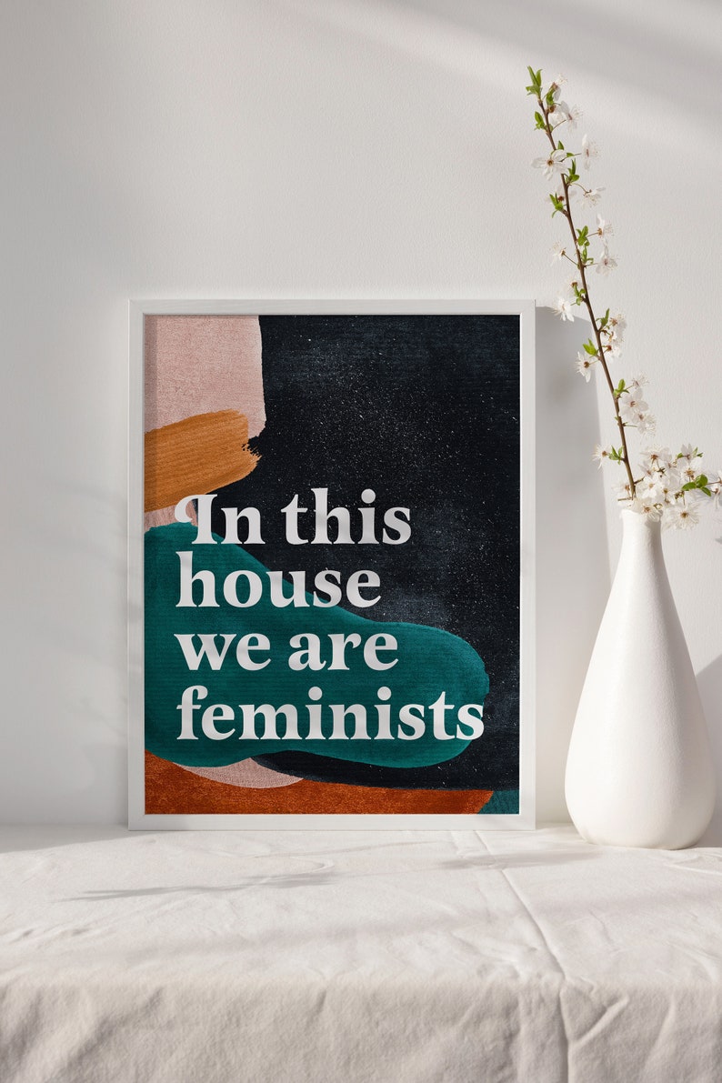 May include: A framed print with an abstract background in shades of pink, orange, brown, and teal. The text "In this house we are feminists" is printed in white on the print.