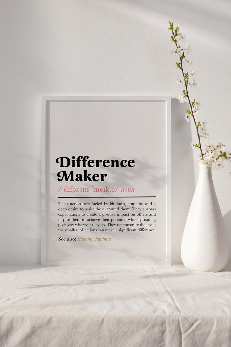 May include: A white framed print with the definition of a "Difference Maker". The definition states that a difference maker is someone whose actions are fueled by kindness, empathy, and a deep desire to assist those around them. The print is on a white surface with a white vase and a branch of white flowers.