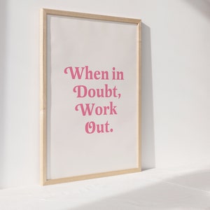 May include: A framed print with the text "When in Doubt, Work Out" in pink lettering. The print is set against a white background and framed in a light wood frame. The artwork is displayed on a white surface.