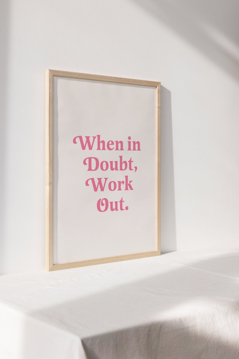 May include: A framed print with the text "When in Doubt, Work Out" in pink lettering. The print is set against a white background and framed in a light wood frame. The artwork is displayed on a white surface.