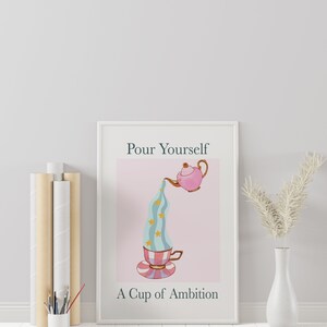 Pu&ograve; includere: Una stampa incorniciata bianca con il testo "Pour Yourself A Cup of Ambition" e una teiera rosa che versa un liquido blu e bianco a righe con stelle dorate in una tazza da t&egrave; con un design a righe rosa e bianco.