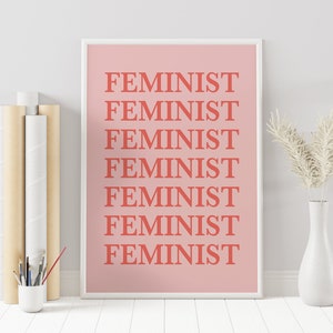 Puede incluir: Un estampado rosa enmarcado con la palabra "FEMINIST" repetida en letras de bloque color coral. El estampado se muestra con un jarr&oacute;n blanco que contiene flores secas y una peque&ntilde;a taza blanca con pinceles.