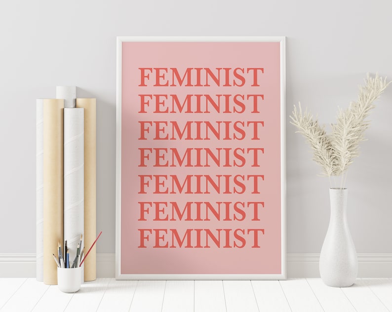 Puede incluir: Un estampado rosa enmarcado con la palabra "FEMINIST" repetida en letras de bloque color coral. El estampado se muestra con un jarr&oacute;n blanco que contiene flores secas y una peque&ntilde;a taza blanca con pinceles.