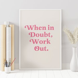 May include: A framed print with the text "When in Doubt, Work Out" in pink lettering. The print is displayed on a light beige background. A white vase with dried flowers and a small white cup with paint brushes are next to the print.