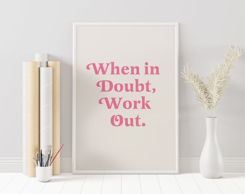 May include: A framed print with the text "When in Doubt, Work Out" in pink lettering. The print is displayed on a light beige background. A white vase with dried flowers and a small white cup with paint brushes are next to the print.