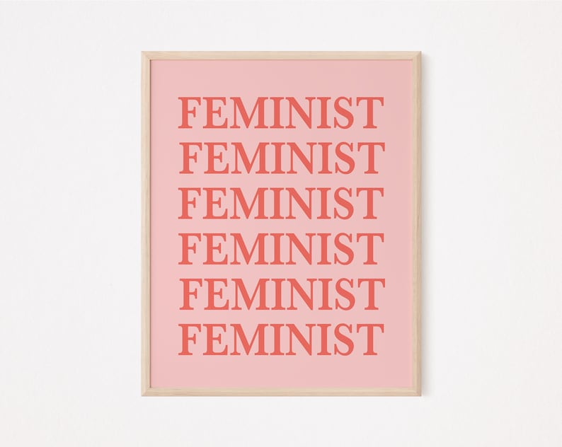 Puede incluir: Un estampado rosa enmarcado con la palabra "FEMINIST" repetida seis veces en una fuente coral audaz. El estampado tiene un fondo rosa claro y un marco de madera simple.