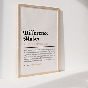 May include: A framed print with a black and white definition of the word "Difference Maker". The definition is in a simple, modern font and includes the phonetic pronunciation of the word. The print is on a white background and has a light wood frame.