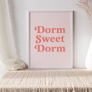 May include: A framed print with the words "Dorm Sweet Dorm" in coral-colored, retro-style lettering on a light pink background. The print is displayed on a beige textured surface, with a white vase and dried plant stem to the right.