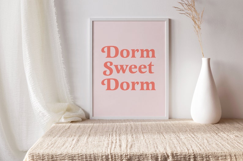 May include: A framed print with the words "Dorm Sweet Dorm" in coral-colored, retro-style lettering on a light pink background. The print is displayed on a beige textured surface, with a white vase and dried plant stem to the right.
