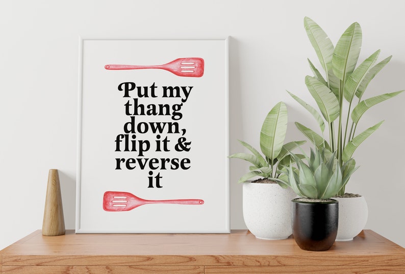 May include: A white framed print with a red spatula on top and bottom. The text on the print reads "Put my thang down, flip it & reverse it".