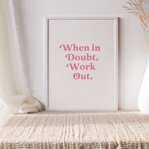 May include: A framed print with the text "When in Doubt, Work Out" in pink lettering. The print is displayed on a beige textured surface, with a white vase holding dried plants to the right. A white curtain is draped on the left.