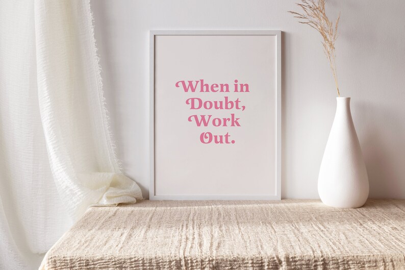 May include: A framed print with the text "When in Doubt, Work Out" in pink lettering. The print is displayed on a beige textured surface, with a white vase holding dried plants to the right. A white curtain is draped on the left.