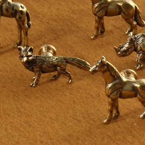 Op de afbeelding: Een set van tien messing dierenknopjes voor lades, waaronder een giraffe, neushoorn, paard, vos en meer. De knopjes zijn allemaal in een gouden afwerking en hebben een gedetailleerd ontwerp.