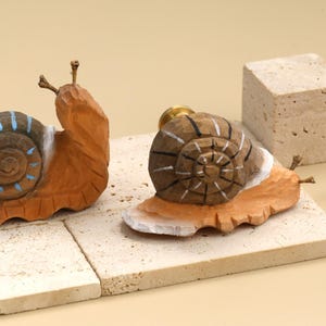 May include: Two hand-carved wooden snail-shaped drawer knobs with brass hardware. The left snail features a brown shell with light blue painted stripes. The right snail has a brown shell with white painted stripes and a white painted base.