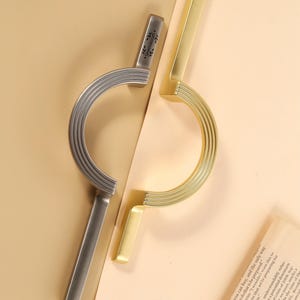 May include: Two cabinet pulls, one silver and one gold, are shown. The silver pull has a curved handle with a ridged design and a rectangular base. The gold pull has a similar design. The brand name "Queeniecraft" is visible.