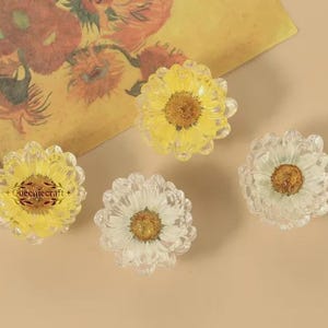 May include: Four daisy-shaped decorative objects with yellow and white petals and a brown center. The objects are made of clear material and are arranged on a beige surface. A piece of art with flowers is in the background.