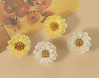 May include: Four daisy-shaped decorative objects with yellow and white petals and a brown center. The objects are made of clear material and are arranged on a beige surface. A piece of art with flowers is in the background.