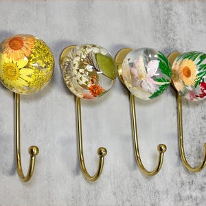 Puede incluir: Cuatro colgadores de pared decorativos con globos de resina transparente que contienen flores secas. Los ganchos tienen un acabado dorado y están montados sobre una superficie gris claro. Cada gancho presenta un arreglo floral diferente, incluyendo flores amarillas, blancas y rosas.