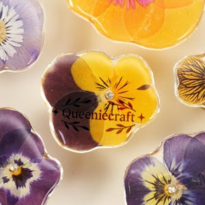 May include: Close-up of several resin flower pendants in various colors, including purple, yellow, and orange. Each pendant features a real pressed flower and a small crystal. The text "Queeniecraft +" is visible on one pendant.