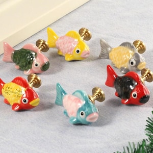 Könnte beinhalten: Eine Sammlung von sechs keramik Fisch-Schrankknöpfen in verschiedenen Farben, darunter Rot, Gelb, Rosa und Blau. Jeder Knopf hat einen messingfarbenen Stiel zur Montage. Einige Knöpfe haben Text.