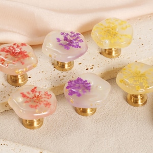 May include: Six decorative cabinet knobs with brass bases. The knobs are made of clear resin, each with a unique floral arrangement in shades of red, purple, and yellow. The knobs are arranged on a neutral surface.