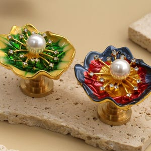 May include: Two decorative floral cabinet knobs. One is green and gold, the other is red and blue. Each knob features a pearl centre and is adorned with small crystals. The knobs are mounted on a gold base.