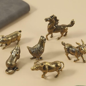 Puede incluir: Una colección de pequeñas figuras de animales de color bronce, que incluyen un cerdo, un ratón, una gallina, un caballo, una cabra y un búfalo de agua. Las figuras son detalladas y tienen una estética vintage. Los animales están dispuestos sobre una superficie beige claro.