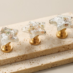 Op de afbeelding: Drie stervormige kastknoppen met doorzichtige harsachtige toppen gevuld met zilveren en gouden accenten. De knoppen hebben een goudkleurige basis en worden getoond op een getextureerd, lichtgekleurd oppervlak. Decoratieve hardware.