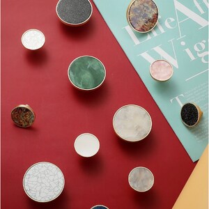 May include: Assortment of 11 round cabinet knobs with various patterns and colors. The knobs are made of brass and feature a variety of materials, including marble, wood, and stone. The knobs are displayed on a red background.