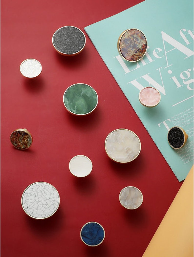 May include: Assortment of 11 round cabinet knobs with various patterns and colors. The knobs are made of brass and feature a variety of materials, including marble, wood, and stone. The knobs are displayed on a red background.