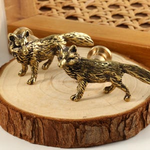 Op de afbeelding: Twee gouden knoppen in de vorm van een vos met een gedetailleerd ontwerp. De knoppen bevinden zich op een houten oppervlak.