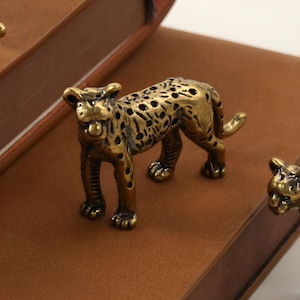 May include: Close-up of three small, detailed brass leopard figurines. The leopards are positioned on a brown leather-bound book. The figurines have a spotted pattern and are in a walking pose. The book has a rounded spine.