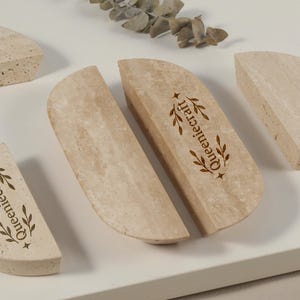 Op de afbeelding: Een verzameling lichtbeige stenen onderzetters met een halfrond ontwerp. De onderzetters hebben een textuuroppervlak en de tekst "Queeniecraft" is op sommige ervan gedrukt. Een takje gedroogde bladeren staat op de achtergrond.