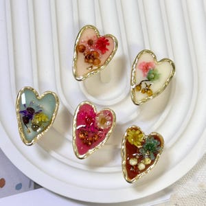 Può includere: Sei anelli in resina a forma di cuore con bordi color oro, ciascuno contenente fiori secchi di vari colori. I colori della resina includono rosa, blu e rosso. Gli anelli sono disposti su una superficie bianca e ondulata.