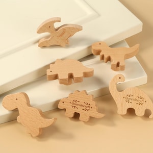 Może przedstawiać: Kolekcja sześciu drewnianych figurek dinozaurów w różnych kształtach i pozach. Dinozaury są jasnobrązowe i mają gładkie wykończenie. Jeden dinozaur ma wygrawerowany napis "Queeniecraft +" z boku.