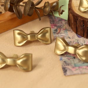 May include: Four gold-coloured bow-shaped drawer pulls. The pulls are made of metal and have a shiny finish.