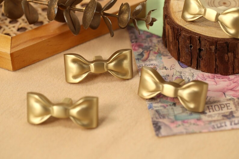 May include: Four gold-coloured bow-shaped drawer pulls. The pulls are made of metal and have a shiny finish.