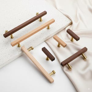 May include: A collection of wooden cabinet pulls in various sizes and shades of brown and natural wood. The pulls have a rectangular design and are mounted on gold-colored hardware. The arrangement includes both long and short pulls, showcasing a range of options for furniture.