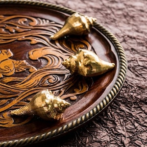Może przedstawiać: Dekoracyjna drewniana taca z misternymi rzeźbami i trzema złotymi muszlami. Taca ma ciemnobrązowe wykończenie ze szczegółowym wzorem fal i brązową obwódką z liny.