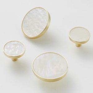 May include: Four round, gold-toned cabinet knobs with white, pearlescent inlays. The knobs are different sizes.