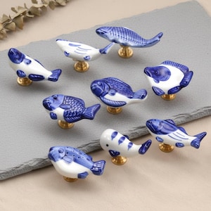 May include: A selection of ceramic fish-shaped cabinet knobs. Each knob is white with blue detailing and a gold-coloured base. The knobs are arranged on a grey rectangular surface, showcasing their decorative design.