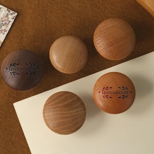 Może przedstawiać: Pięć drewnianych gałek do szafek w różnych odcieniach brązu. Dwie gałki mają nadruk "+ Queeniecraft +". Gałki ułożone są na brązowej powierzchni z kwiatową kartą i białą kartą.